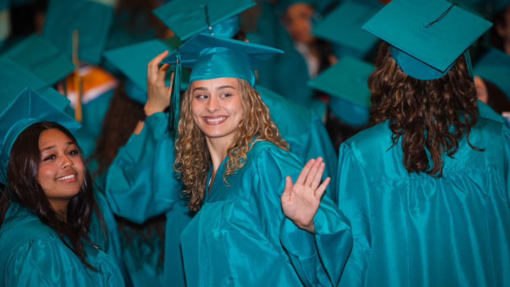 Group of graduates in teal regalia with one woman looking back, smiling, and waving