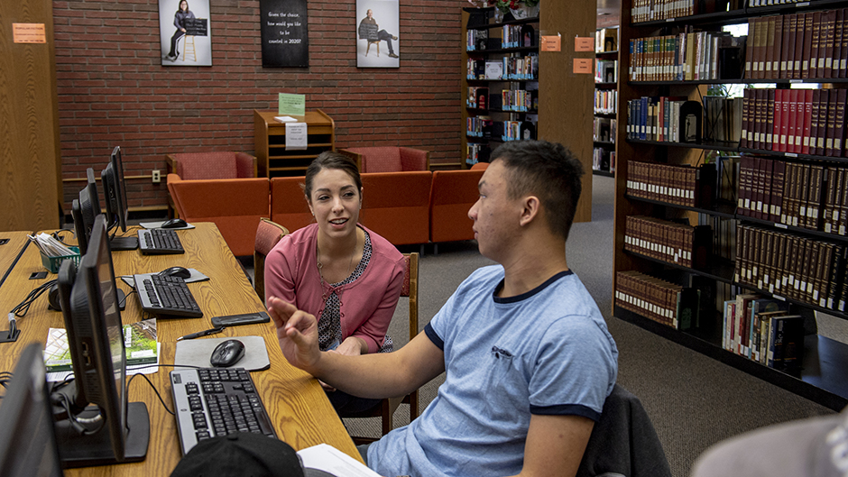 A male student and female faculty member talking to each other while sitting at computers in the library